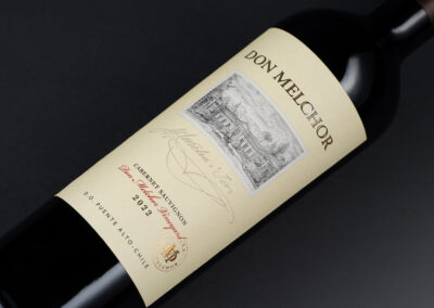 Botella de vino tinto con etiqueta clara. La etiqueta presenta el nombre "Don Melchor" en letras grandes y doradas, junto con la variedad "Cabernet Sauvignon" y el año "2022". En la parte superior, hay una ilustración en blanco y negro de una casa rodeada de árboles. En la parte inferior, se indica "D.O. Puente Alto-Chile" y un sello dorado con un emblema. El fondo es negro, lo que resalta la botella.