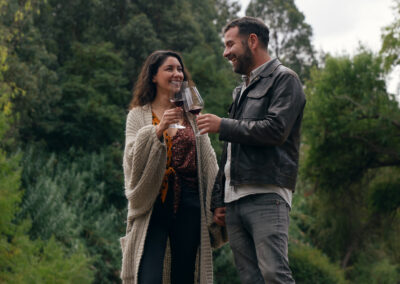 Una pareja sonriente sostiene copas de vino en un entorno natural. La mujer lleva un suéter largo y una blusa con estampado, mientras que el hombre viste una chaqueta de cuero y jeans. Ambos parecen disfrutar de un momento juntos, rodeados de árboles y vegetación. El cielo está nublado, lo que sugiere un ambiente fresco.