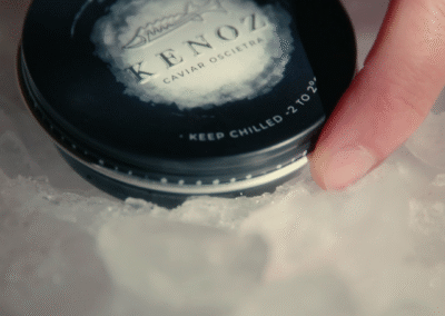 Un tarro de caviar está colocado sobre hielo. El tarro es de color negro con una tapa que tiene el nombre "KENOZ" y la frase "CAVIAR OSCIETRA". Se puede ver una mano que toca suavemente el tarro. El hielo que lo rodea es de forma irregular y brilla, sugiriendo que está bien frío.